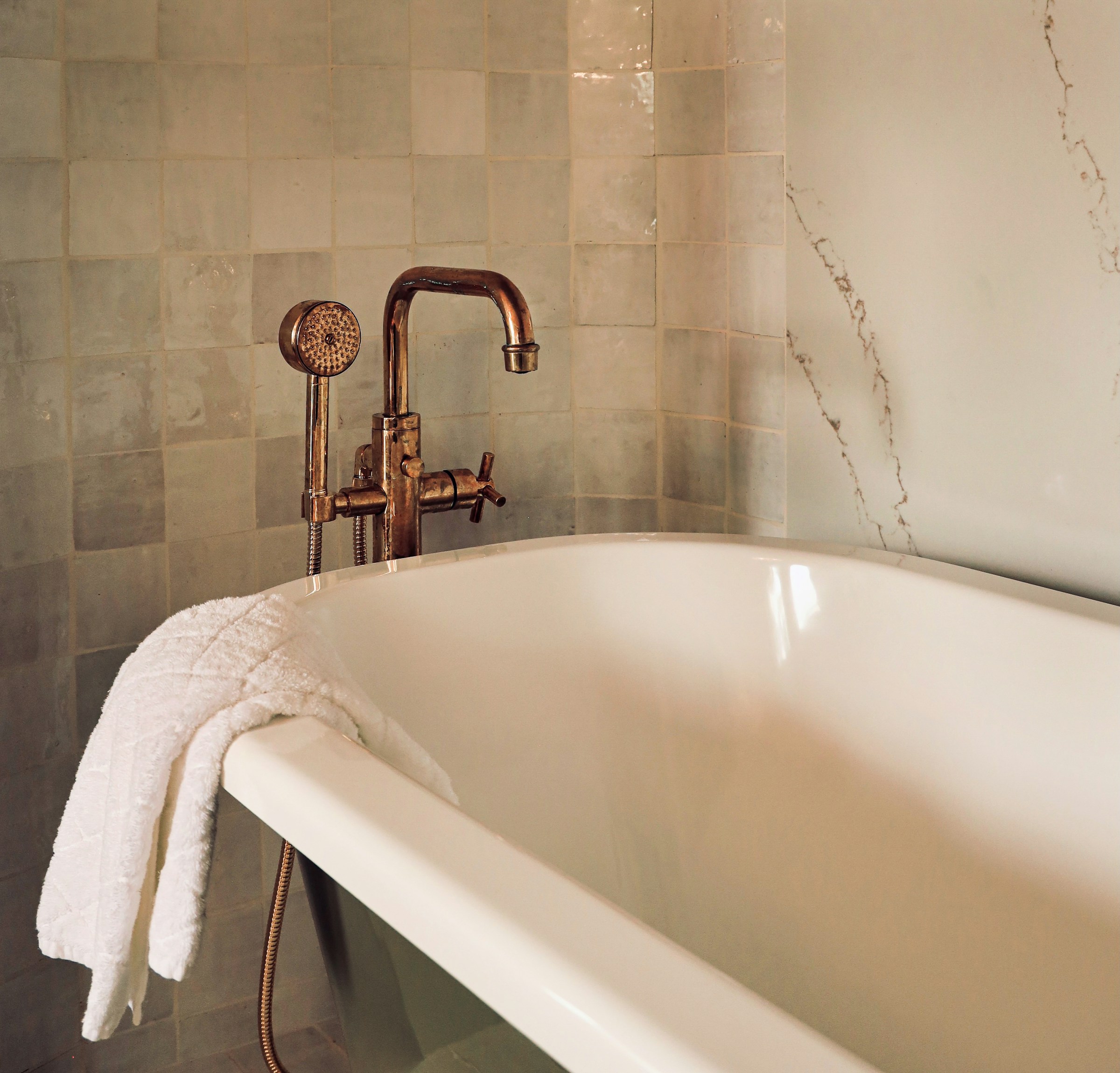A freestanding bathtub with a floor-mounted tap and handheld shower head against a tiled wall